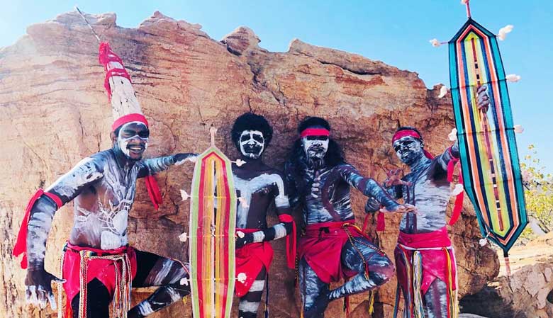 Indigenous Rangers and Traditional Owners painted with body art standing in front of a rock preparing to dance for tourists