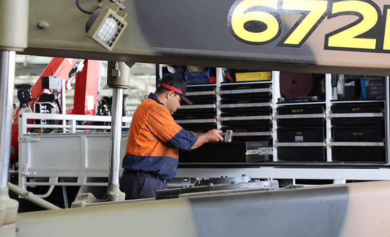 Man in work wear stands next to metal shelving with tools. Behind him is the bed of a truck and behind that, heavy work equipment.