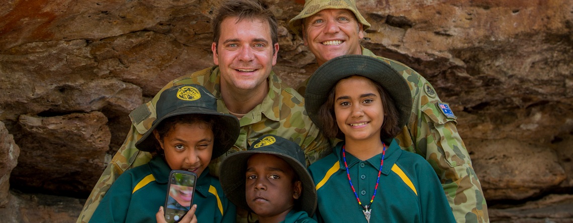 Two male members of the Australian Army, in uniform, with three Indigenous girls, standing in front of some Indigenous rock art