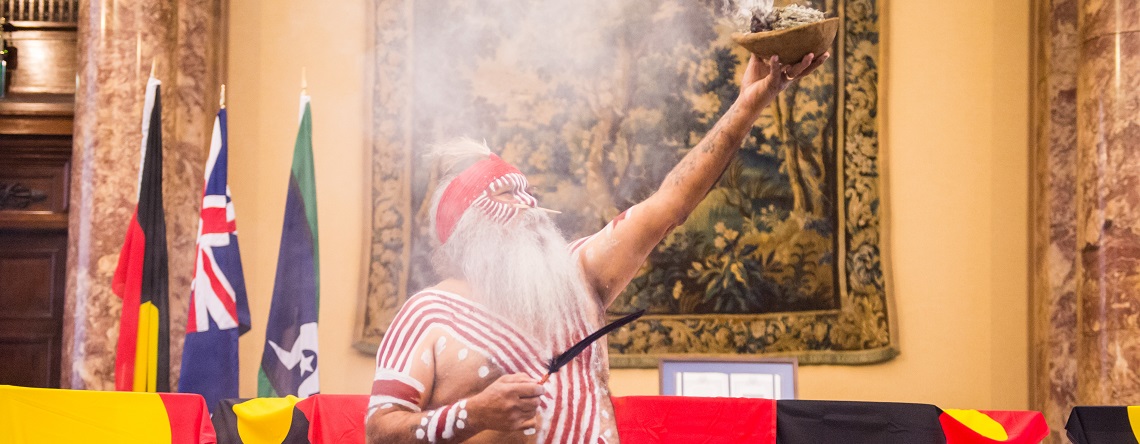 Indigenous elder dressed in traditional dress, holding a smoking bowl up in the air, with ATSI and Australian flags in the background