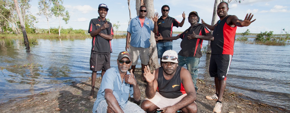 Members of the Arnhem Land Progress Aboriginaol Corporation standing or crouching on the banks of some water, waving to camera