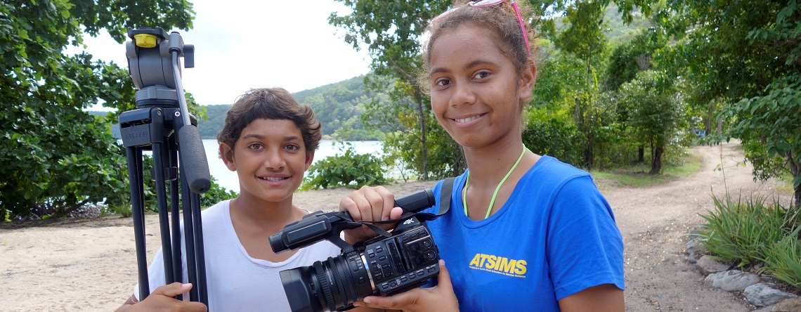 Two Indigenous students holding a camera and tripod with coastline behind them