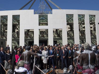 A large group of polititians, community members and Indigenous community members in traditional attire, standing in front of Parliament House