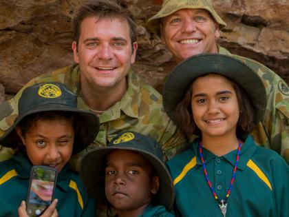 Two male members of the Australian Army, in uniform, with three Indigenous girls, standing in front of some Indigenous rock art