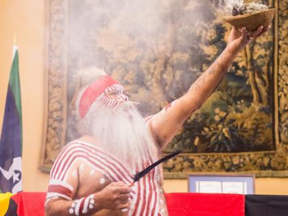 Indigenous elder dressed in traditional dress, holding a smoking bowl up in the air, with ATSI and Australian flags in the background