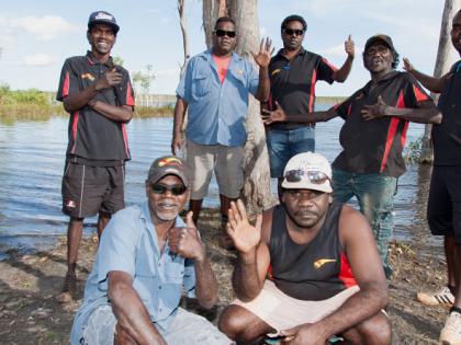 Members of the Arnhem Land Progress Aboriginaol Corporation standing or crouching on the banks of some water, waving to camera