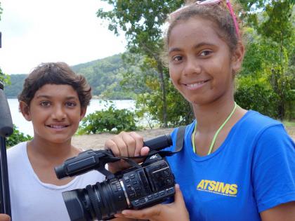 Two Indigenous students holding a camera and tripod with coastline behind them