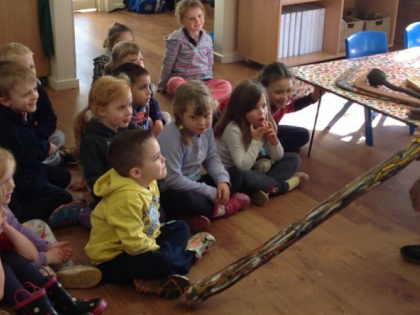 Preschool students sitting on the floor of their classroom, listening to an Indigenous man playing the didgeridoo
