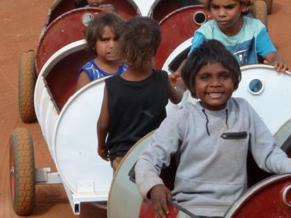 This photo captures Indigenous school students enjoying a fun ride to school at Punmu in the Pilbara in Western Australia. They are in 44 gallon drums being towed along a red dirt road. 