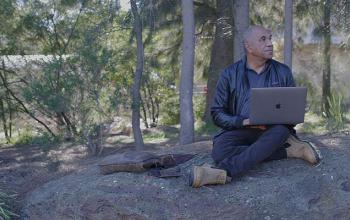A man sits cross-legged under some trees in an Australian bushland setting. There is a body of water behind him, which can be seen between the trees. An open laptop computer rests on one knee.