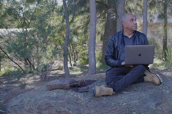 A man sits cross-legged under some trees in an Australian bushland setting. There is a body of water behind him, which can be seen between the trees. An open laptop computer rests on one knee.