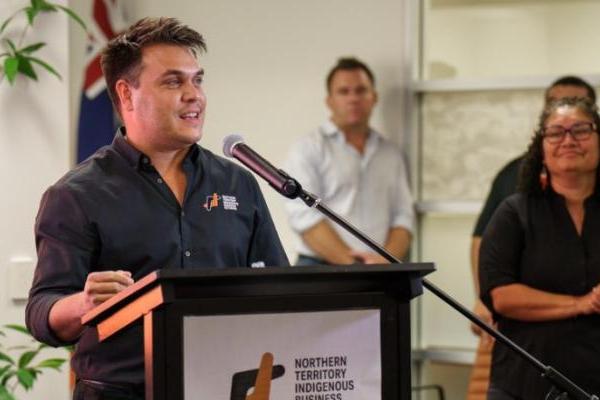 One person, dressed in black shirt with NTIBN logo is the focus of this image.  They are speaking into a microphone behind a lectern which displays a sign showing a logo which reads “Northern Territory Indigenous Business Network”.  The person is addressing a room and is smiling.   Three people appear in the background of the photo and are looking at the speaker.