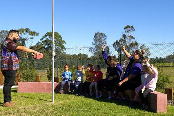 Group of children and adults sit on a low wall. In front is a grassed area on which is a pole. On the other side of the pole stands an Aboriginal man. In the background is a fence and trees and more grass.