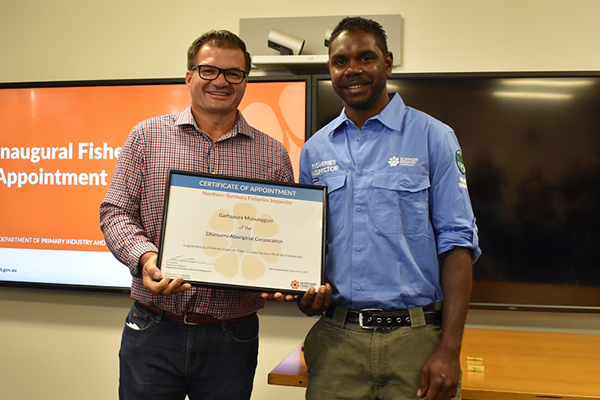 Two men stand side by side and hold between them a large framed certificate. The man on the left wears a checked shirt and dark trousers. The man at right wears a blue shirt and green trousers. They stand in a room with two large TV screens in the background.