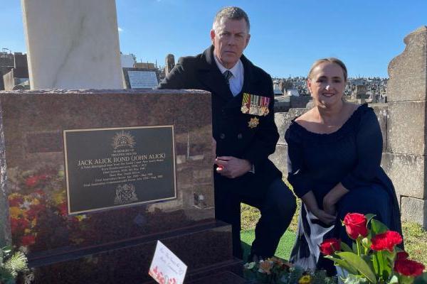 A man wearing a tie and black coat with military medals, and a woman in a black dress kneel next to a grave site. The grave is adorned with bouquets of flowers and has a plaque identifying it as that of Jack Alick Bond, a veteran of the Boer War.