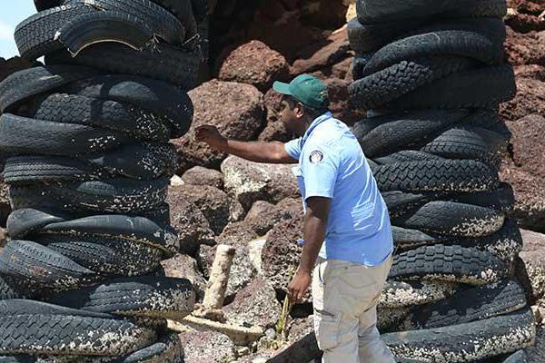 A man under a bridge and surrounded by rocks is standing between two towers built of used car tyres. The man is leaning over to the one of the tower of tyres and is holding a yellow tool in his right hand. The man is dressed in a blue shirt and wearing a green baseball cap.
