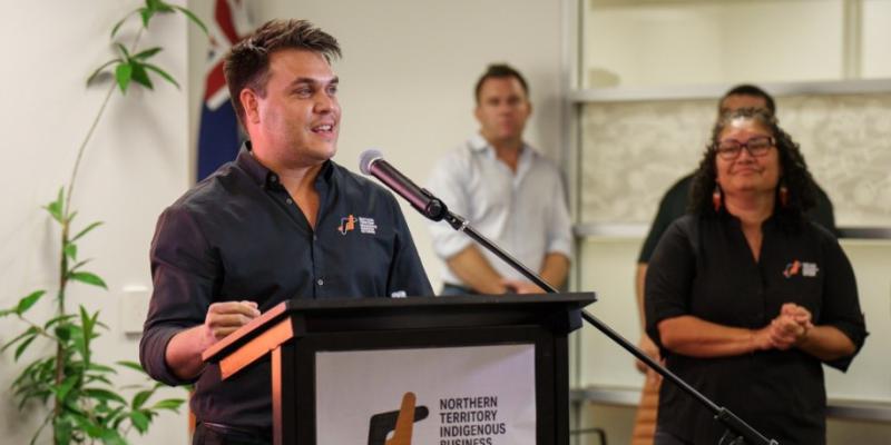 One person, dressed in black shirt with NTIBN logo is the focus of this image.  They are speaking into a microphone behind a lectern which displays a sign showing a logo which reads “Northern Territory Indigenous Business Network”.  The person is addressing a room and is smiling.   Three people appear in the background of the photo and are looking at the speaker.