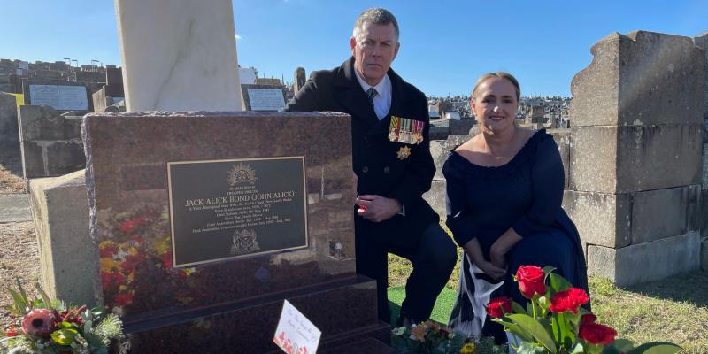 A man wearing a tie and black coat with military medals, and a woman in a black dress kneel next to a grave site. The grave is adorned with bouquets of flowers and has a plaque identifying it as that of Jack Alick Bond, a veteran of the Boer War.