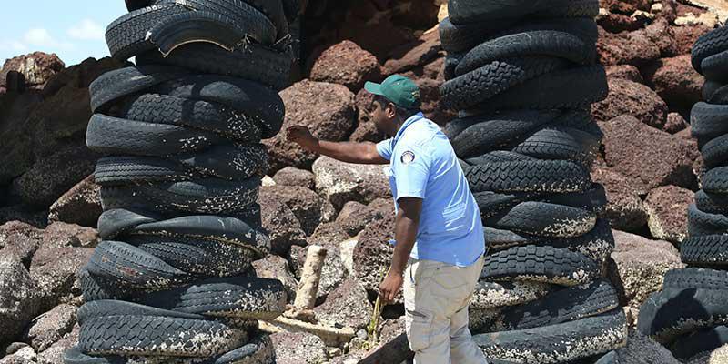 A man under a bridge and surrounded by rocks is standing between two towers built of used car tyres. The man is leaning over to the one of the tower of tyres and is holding a yellow tool in his right hand. The man is dressed in a blue shirt and wearing a green baseball cap.