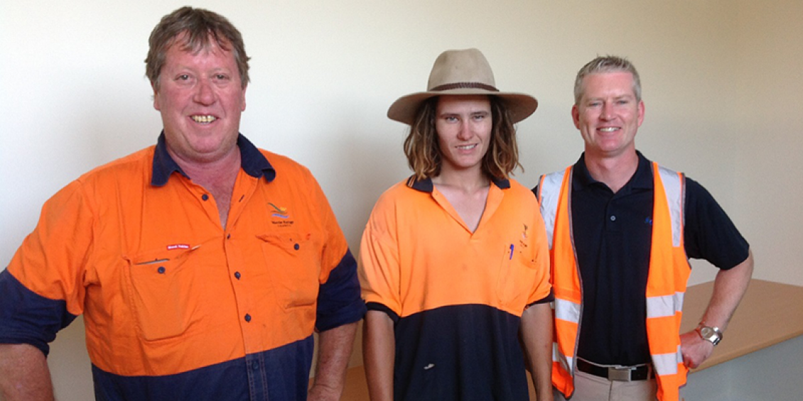 Three men stand wearing predominantly orange work wear. The one in the middle is younger and wears a broad brimmed hat.