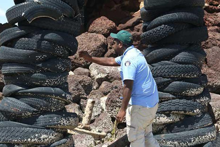 A man under a bridge and surrounded by rocks is standing between two towers built of used car tyres. The man is leaning over to the one of the tower of tyres and is holding a yellow tool in his right hand. The man is dressed in a blue shirt and wearing a green baseball cap.